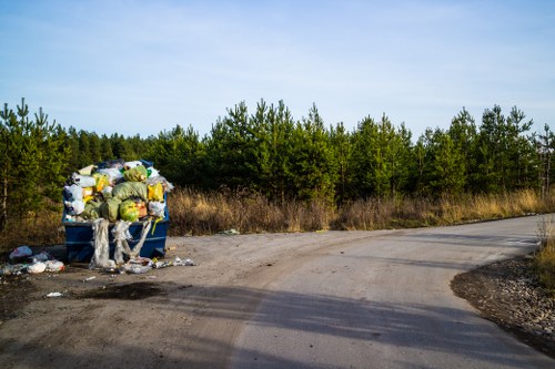 A collection of overflowing rubbish bags and discarded waste materials piled beside a gravel or dirt road on the edge of a wooded area, with green trees and a clear sky in the background. The waste includes plastic bags, cardboard, and other miscellaneous trash, some of which has spilled onto the ground. The scene highlights an unmanaged litter accumulation close to a countryside or suburban environment, reflecting a need for professional rubbish removal services such as those offered by Office Clearance Raynes Park, which specialises in clearing waste from residential or commercial sites in the local area near Raynes Park and postal code SW20.