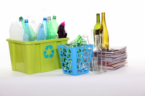 The image displays two containers filled with various glass bottles and cutlery, placed on a plain white surface. The larger container on the left is bright green with a recycling symbol printed on its side, containing several plastic bottles in different colours including white, green, and blue, with some featuring screw caps. The smaller container on the right is blue with cutouts, holding more glass bottles, mostly clear or green, in varied sizes. To the right of the containers, there is a neatly stacked pile of newspapers or magazines, and several empty glass tumblers and bottles, some with labels removed, are arranged beside them. The background is plain white, providing a clear view of the items, which are part of a glass and paper waste collection setup often handled during rubbish removal services, such as those provided by Office Clearance Raynes Park, in a clean and organized environment. The scene emphasizes the separation of recyclable materials and general waste collection in a local setting.