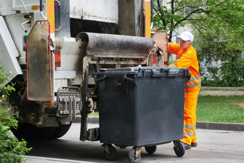 A waste collection worker dressed in a high-visibility orange uniform with reflective stripes and a white cap is operating a rubbish bin lift on a street within an urban area near Raynes Park. The worker is standing on a paved surface beside a large black wheeled rubbish bin, which is positioned directly beneath the mechanical arm of the rubbish collection truck. The truck, featuring a metallic and painted exterior with orange safety accents and visible mechanical components, is actively engaged in emptying the bin. The background includes trees and a grassy verge, indicating a residential or park-adjacent street environment. The scene is set during daylight hours, and the overall atmosphere is organized and professional, fitting within the context of rubbish removal services in the Raynes Park area of London.