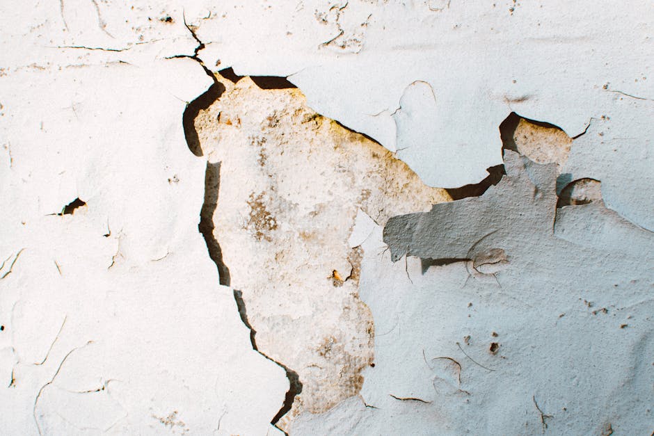Close-up photograph of a wall with peeling paint revealing underlying layers. The top layer of paint is light blue, chipped and flaking off in irregular patches, exposing a layer of beige or light bro