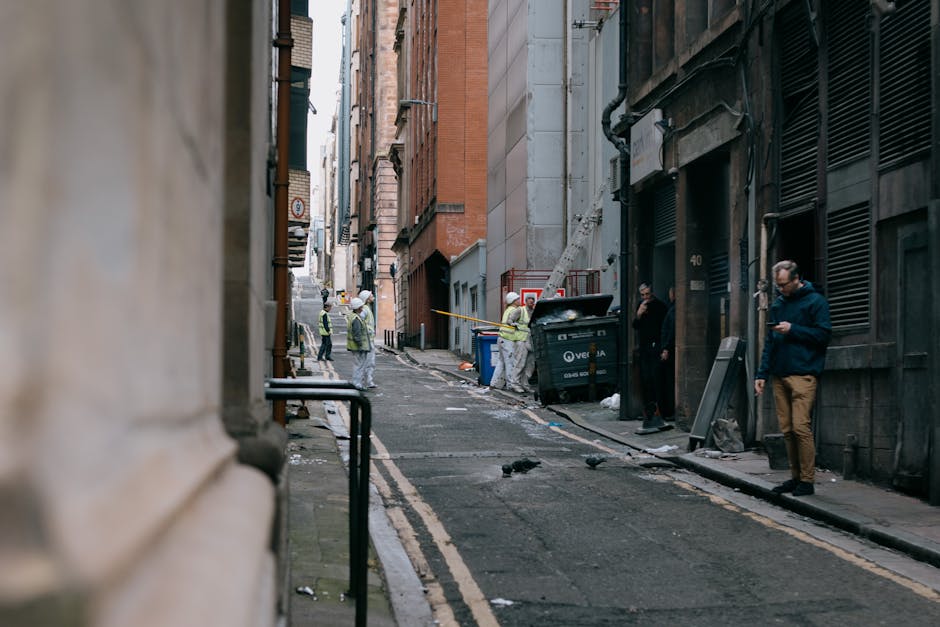 A worn, dirty fabric trash bag filled with mixed waste, including a plastic container visible at the top, is placed on a narrow urban street sidewalk next to a weathered wall with metal bars on the wi