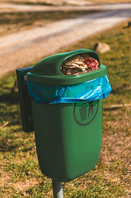 A close-up of a person's hand placing a used yellow and blue lithium-ion battery into a green recycling bin designated for batteries and electronic waste. The bin has a rectangular opening with a blac